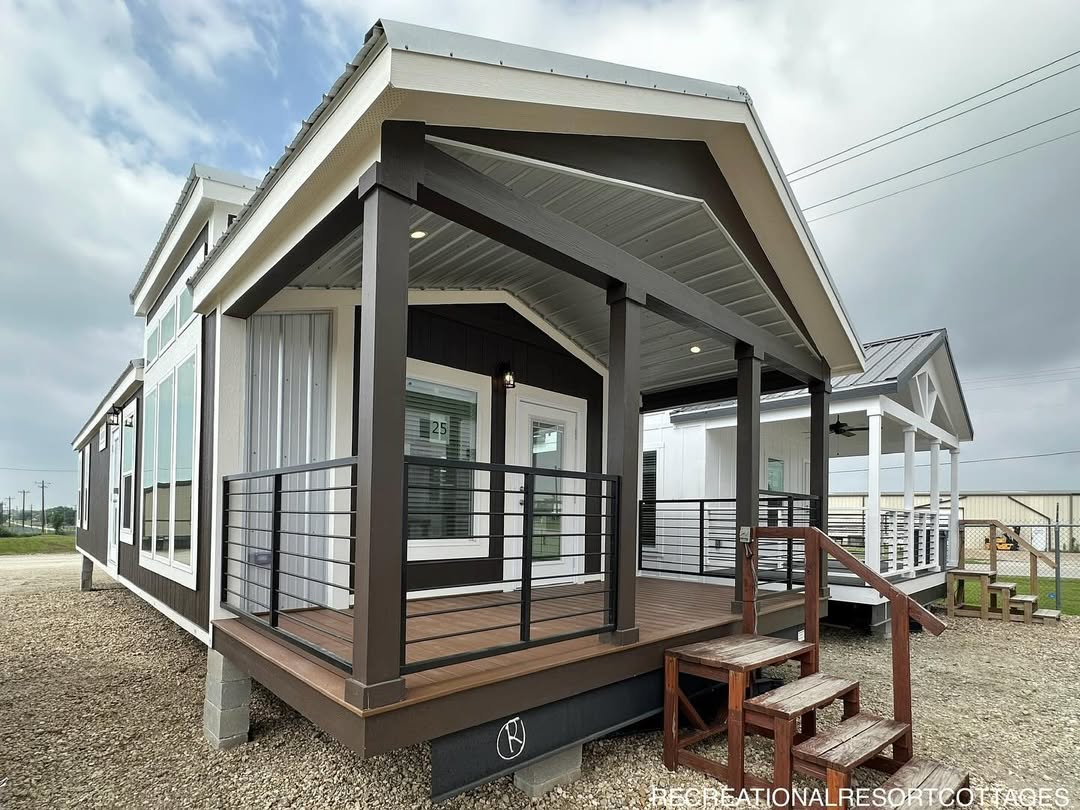 Small modern cabin with a covered porch, wooden steps, and black railings. The exterior is dark with white trim. Cloudy sky in the background.