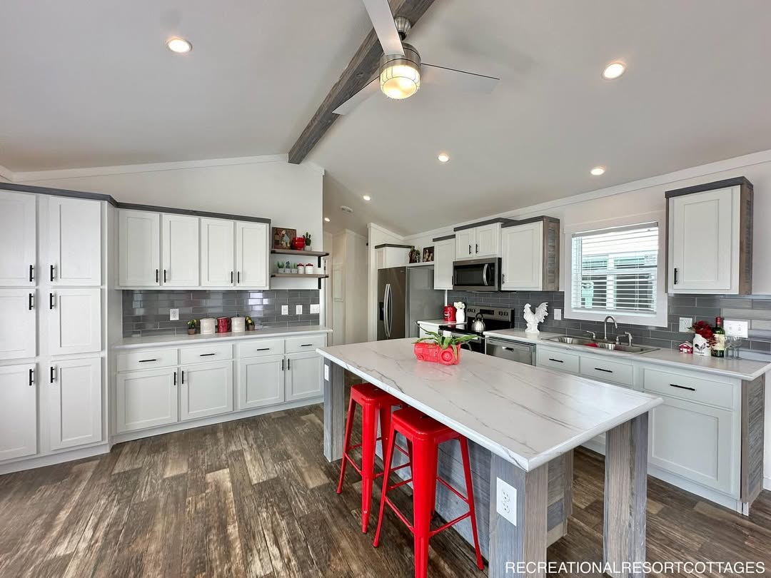 Modern kitchen with white cabinets, gray countertops, and wood flooring. A large island with red stools and a ceiling fan add vibrant touches.