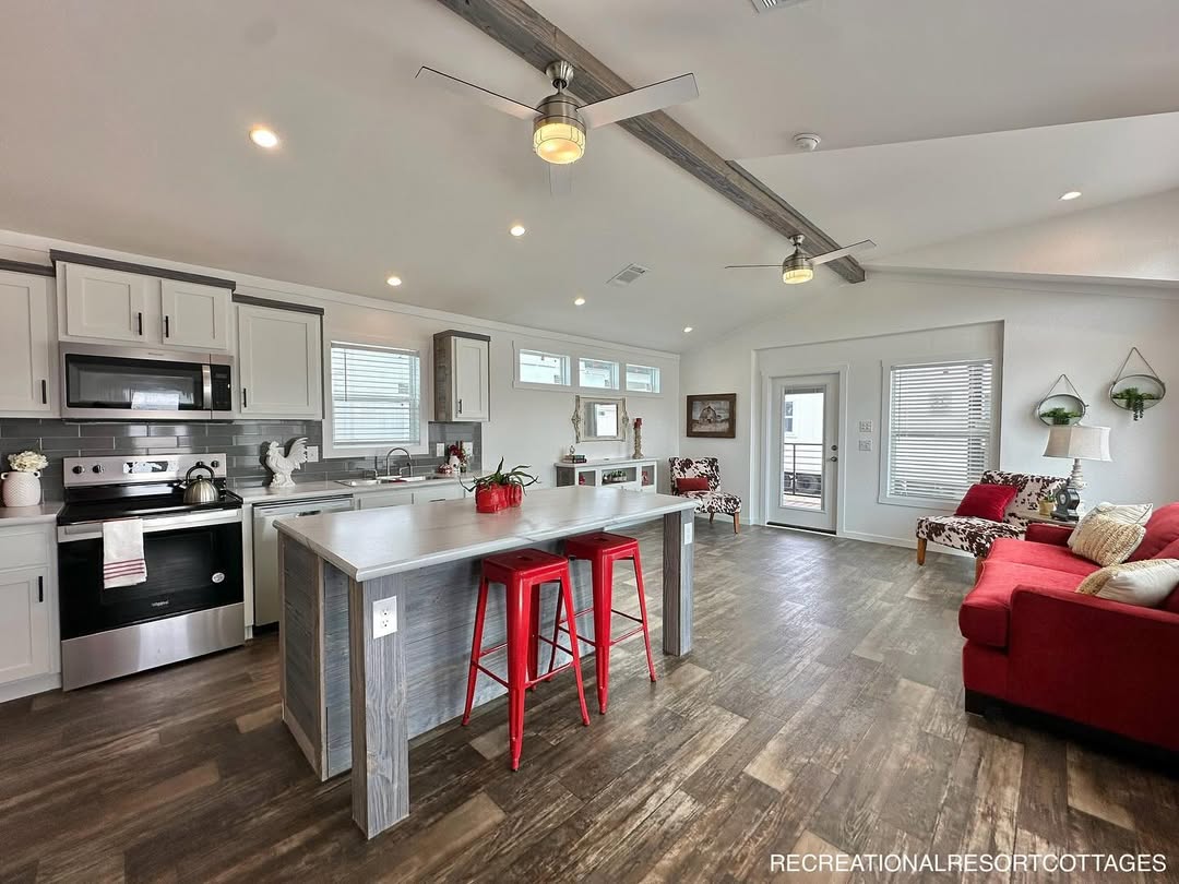 Modern, bright kitchen and living area with vaulted ceiling. Features wood floors, gray cabinets, and stainless steel appliances. Red stools and sofa add pops of color, while large windows bring in natural light. Cozy, inviting atmosphere.