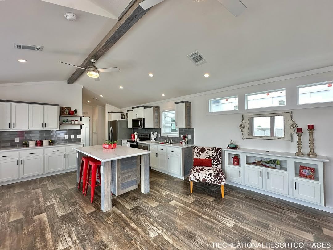 Spacious modern kitchen with white cabinets, gray backsplash, and wood accents. Features a central island with red stools, and a cozy patterned chair.