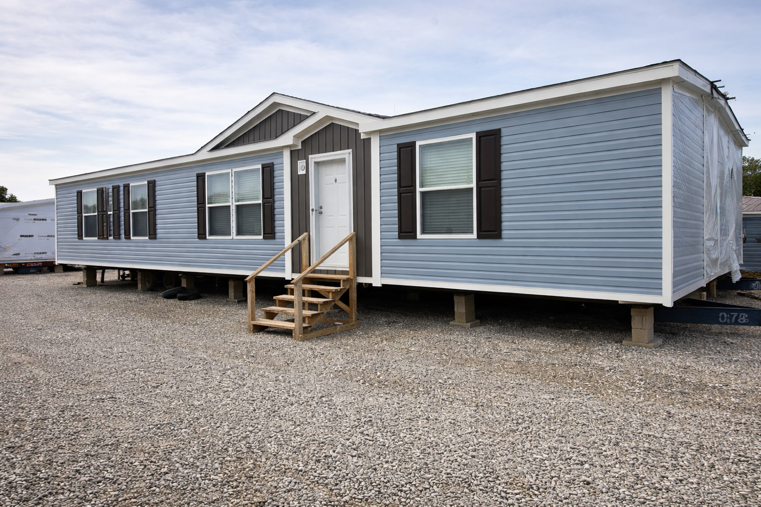 A light blue mobile home with white trim and dark shutters sits elevated on concrete blocks over a gravel lot. Wooden steps lead to the front door.