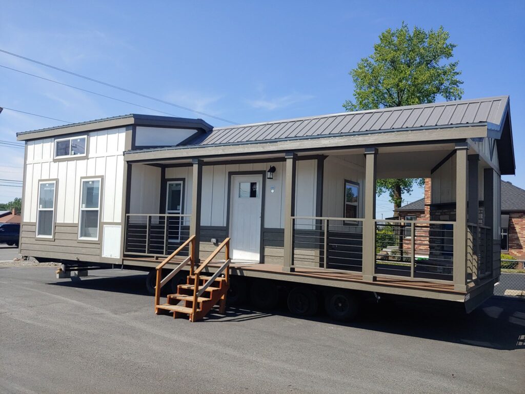 A modern tiny house with a pitched metal roof and large windows, set on wheels in a parking lot. It has a porch with railings and wooden steps.