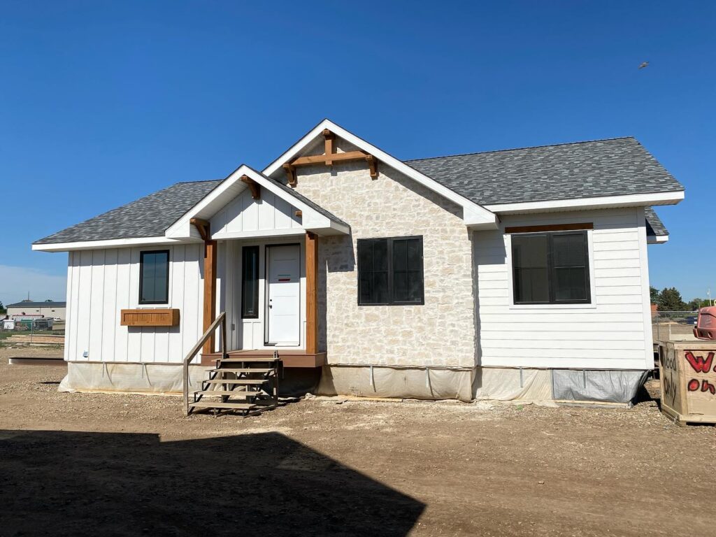 A newly constructed single-story house with white siding and stone accents. It has a gray shingled roof and small front porch on a dirt lot under clear blue sky.