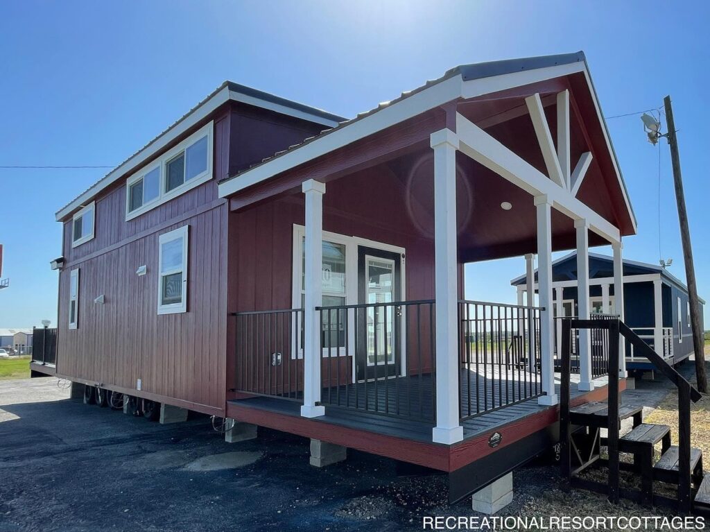 Red tiny house with white trim and a covered porch under a clear blue sky. The structure is elevated, adding depth and accessibility steps. Cozy and inviting.