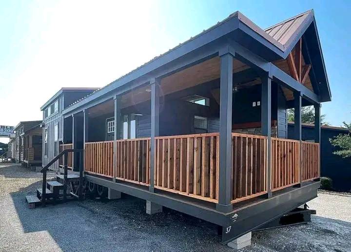 A modern tiny house with a dark exterior and a large wooden porch stands on a gravel lot. The pitched roof and wooden railings offer a rustic charm.