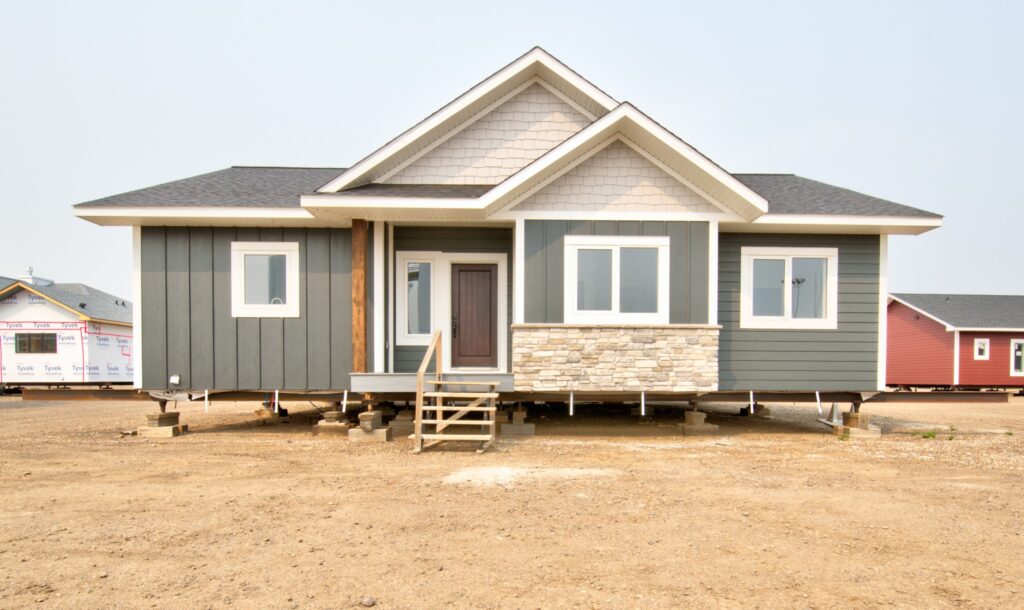 A newly constructed modular house on a dirt lot, featuring gray siding, white trim, and stone accents under a partly cloudy sky, conveying a sense of completion.