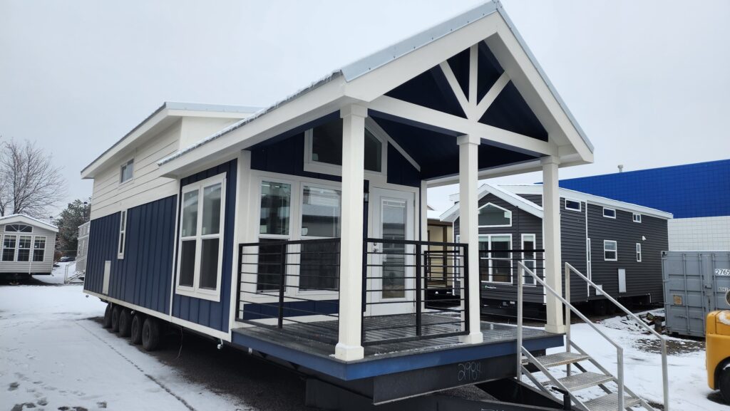Modern tiny house with a blue and white exterior on a snowy lot. Features large windows, a small porch with black railings, and a gabled roof.