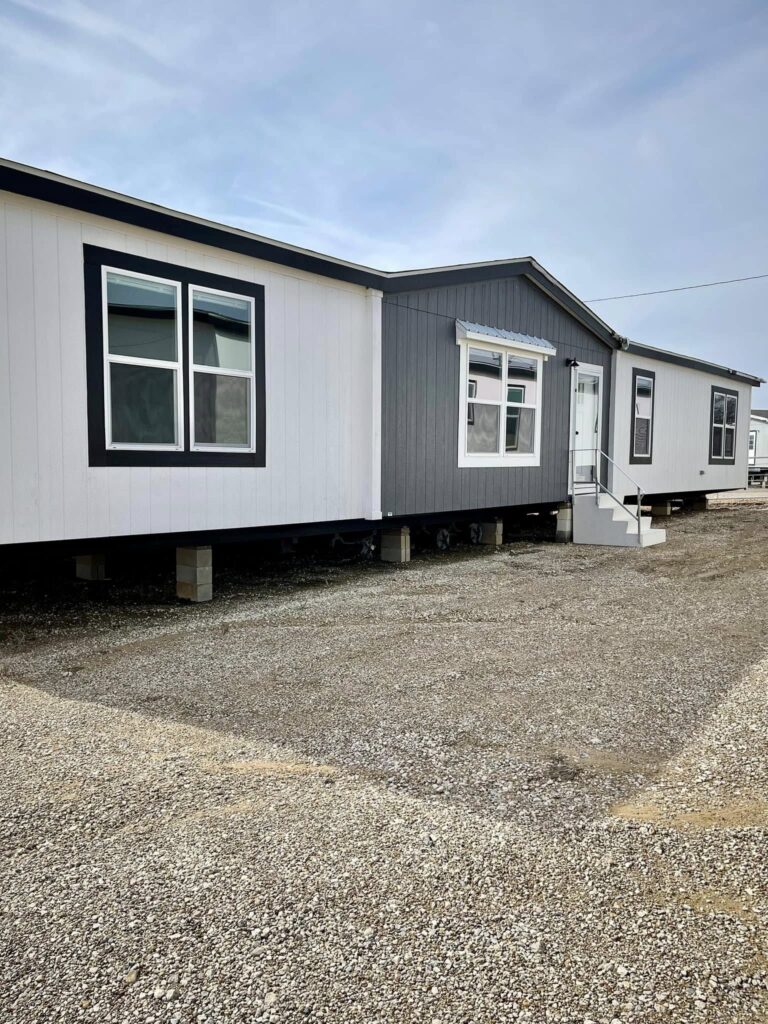 A modern double-wide mobile home sits on a gravel lot, featuring white siding and a gray accent wall. Large windows and a small porch add a welcoming feel.