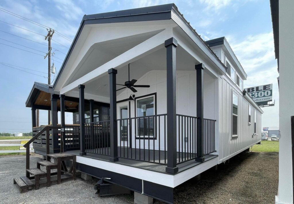 Modern white mobile home with black trim, featuring a covered porch with a ceiling fan and railing. Stairs lead up, set against a clear blue sky.