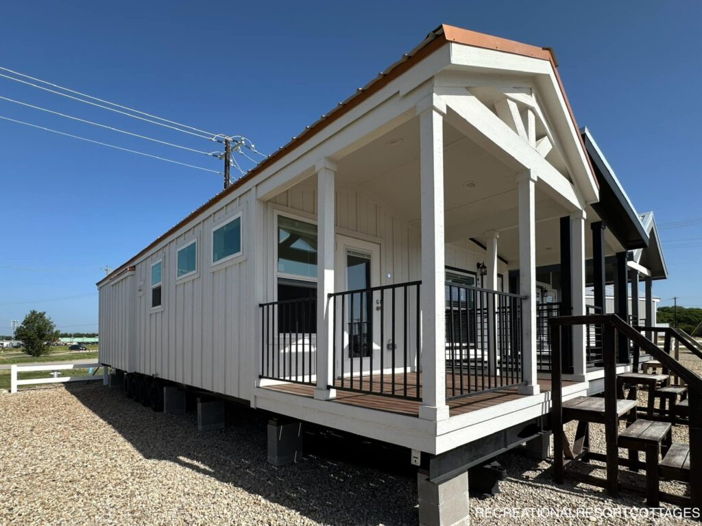 A modern tiny house with a white exterior and a pitched roof sits elevated on a gravel lot. It features a front porch with black railings, under a clear blue sky.