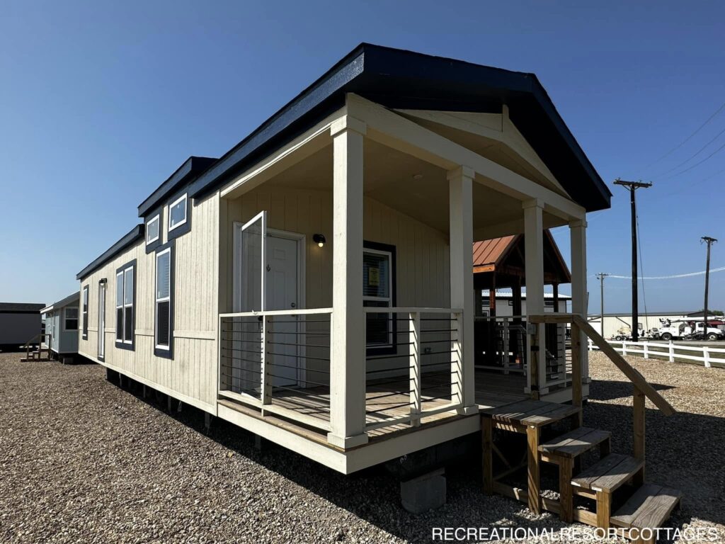 Alt text: A beige modular home with a dark blue roof is situated on a gravel lot under a clear blue sky. The home features a small wooden porch with railings and steps leading up to the entrance. In the background, there are other similar homes and utility poles. The atmosphere appears calm and quiet.