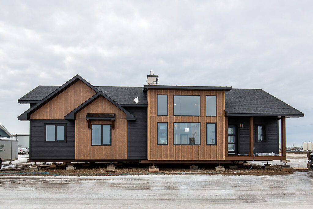 Modern single-story house with a mix of dark wood and black siding. Large windows enhance brightness. Snowy ground and overcast sky create a cold ambiance.