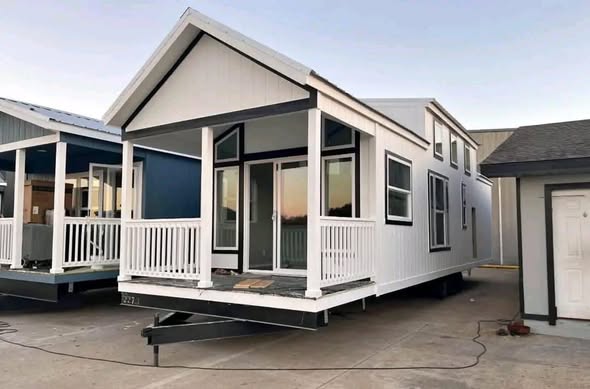 A modern tiny house with white siding and black trim sits on a trailer. It features large windows, a small porch, and a pitched roof, exuding a cozy feel.