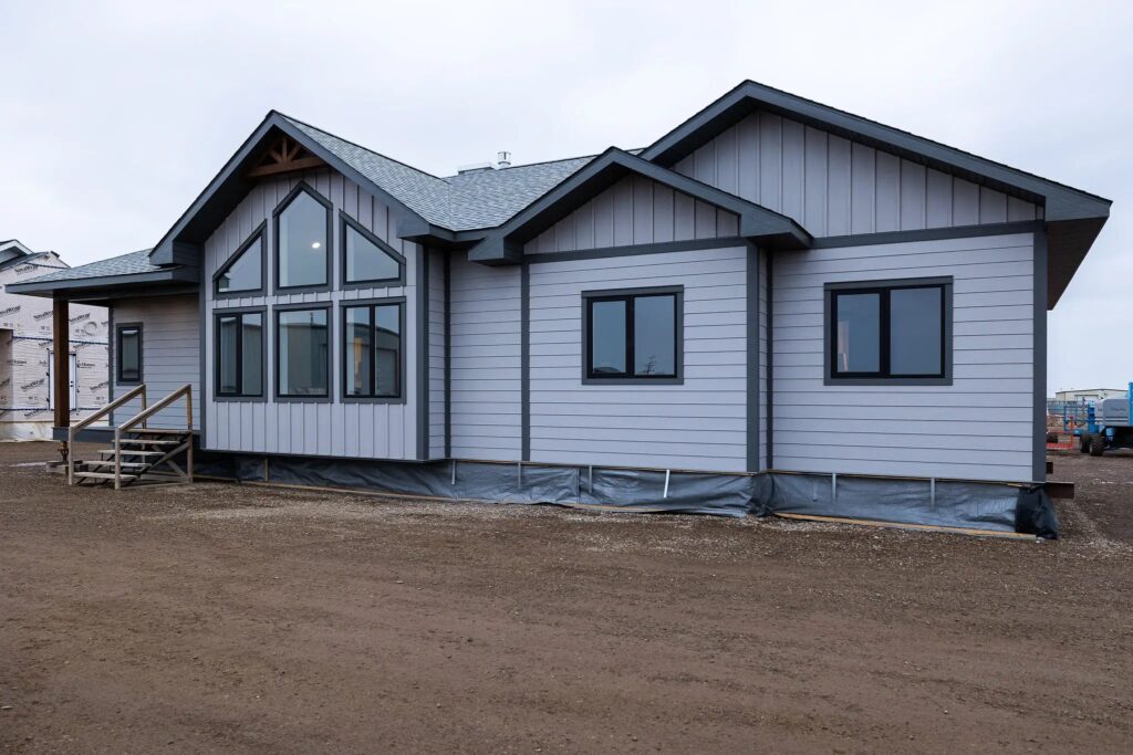Newly constructed house with light gray siding and black-trimmed windows under a cloudy sky. The yard is barren, indicating early stages of landscaping.