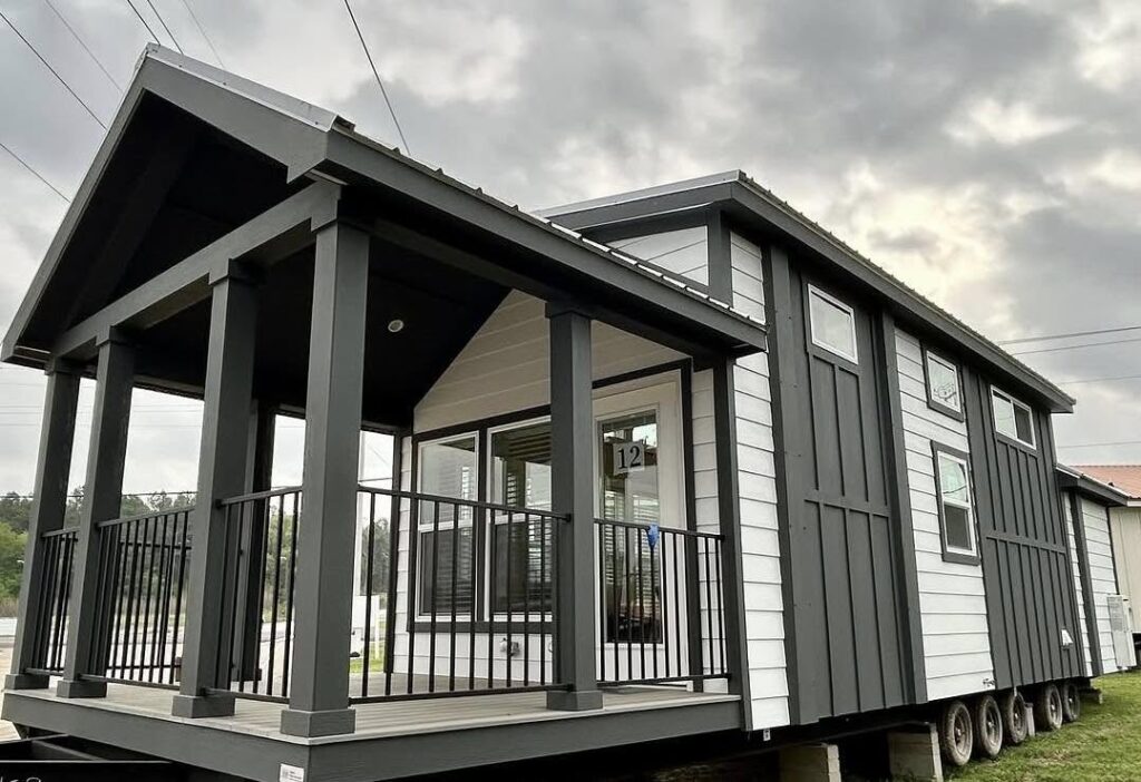 Modern tiny home with gray and white siding on a trailer base. It features a covered porch with black railings under a cloudy sky.
