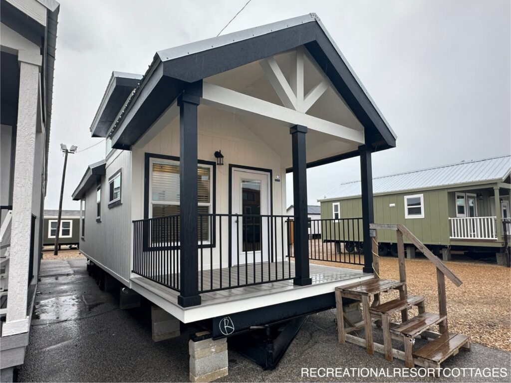 Compact modern tiny house with a gabled roof, white siding, black trim, and a covered porch. Wooden steps lead up to the porch, creating a welcoming vibe.