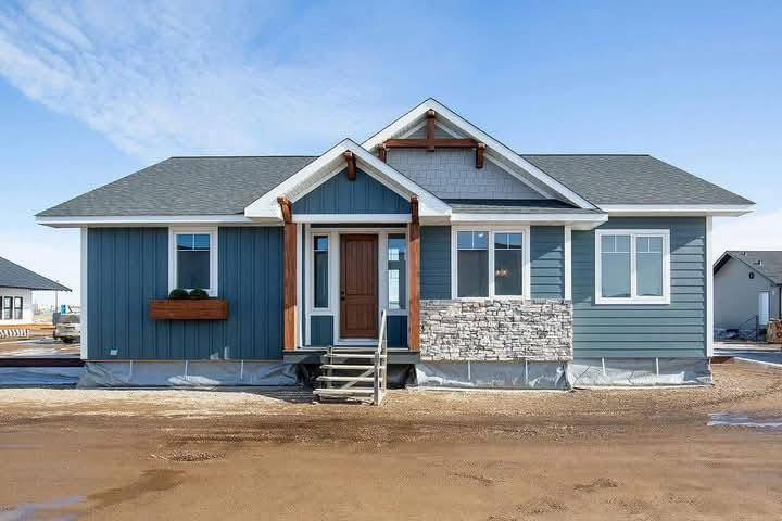 Single-story blue house with stone accents, wood trim, and a peaked roof under a bright blue sky. Entryway features a small porch and steps.