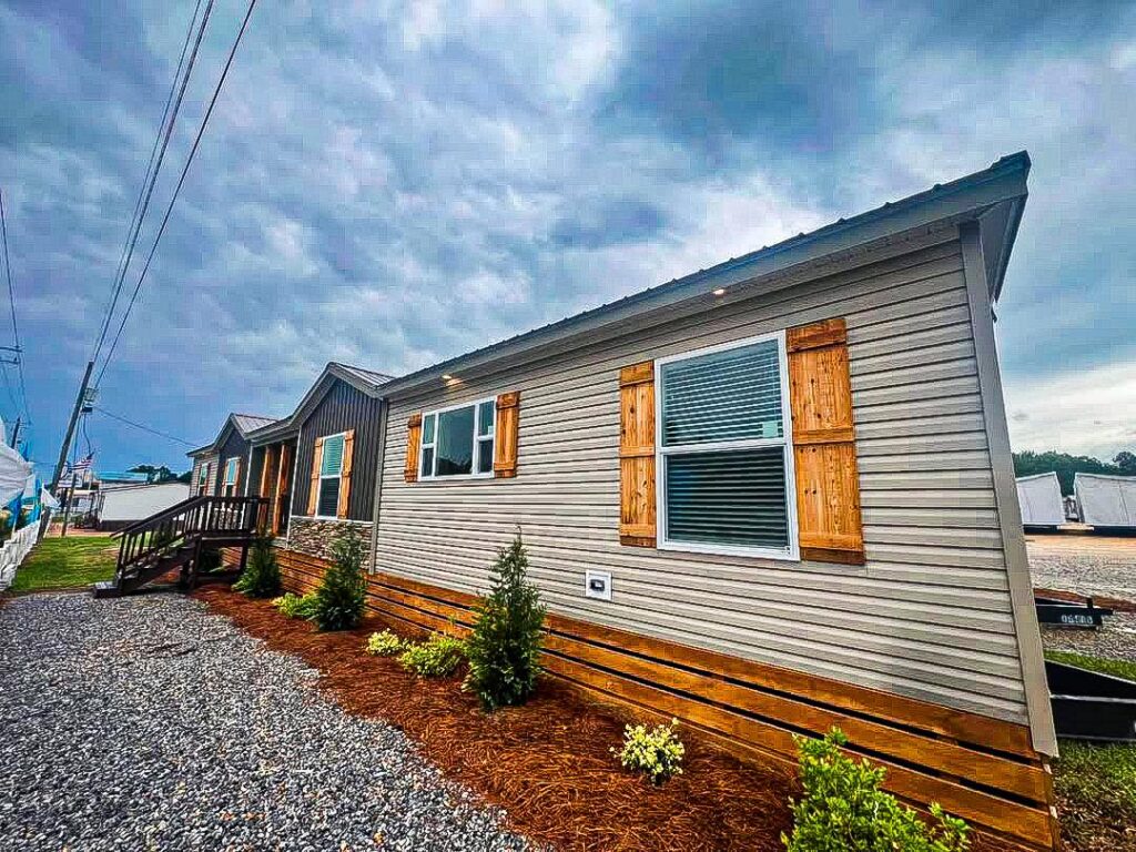 Manufactured home with wood shutters and siding, surrounded by small bushes on a gravel pathway. Overcast sky creates a calm, subdued atmosphere.