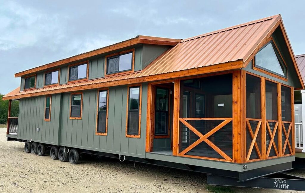 A modern tiny house on wheels with dark green siding, multiple large windows, and a prominent wooden porch with a steep, metal gabled roof.