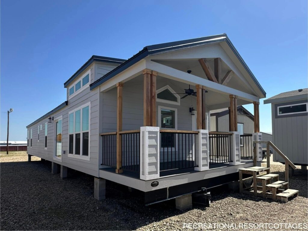 A modern tiny home with a gray exterior and white trim stands under a clear blue sky. The home features a covered wooden porch with steps, large windows, and a pitched roof, conveying a cozy and inviting feel.