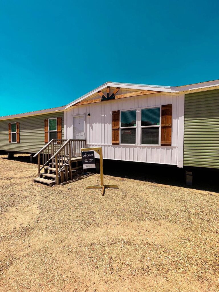 A single-story manufactured home with light green siding and wood accents is set against a clear blue sky. A small sign is in front by the wooden steps.