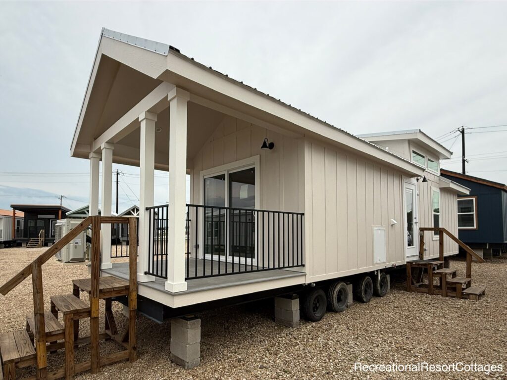 Compact, beige tiny house with a gabled roof and front porch on wheels. Wooden stairs lead to the entrance. Clear sky and gravel ground surround it.