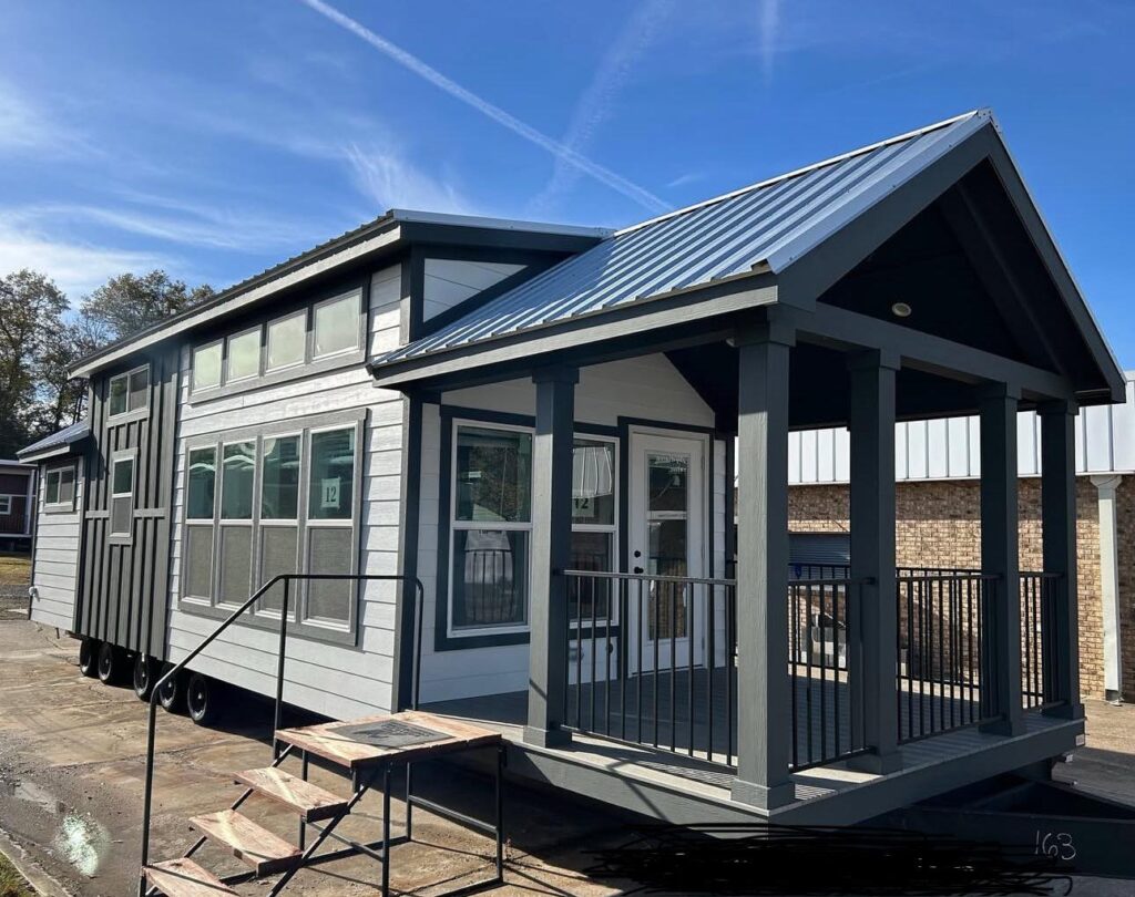 A modern tiny house on wheels with a gray exterior and a blue metal roof. It features a covered porch with railings and large windows, under a clear blue sky.