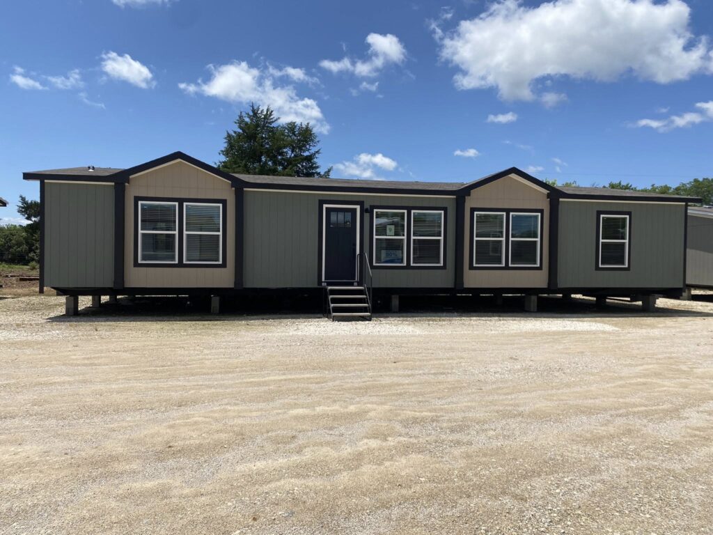 A large beige and gray mobile home with a black door and several windows sits on a gravel lot under a bright blue sky with scattered clouds.