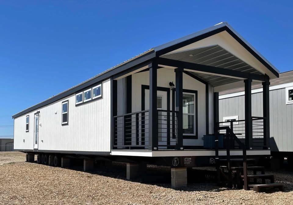 A modern white manufactured home with black trim sits elevated on a gravel lot. It features a covered porch with steps, under a clear blue sky.