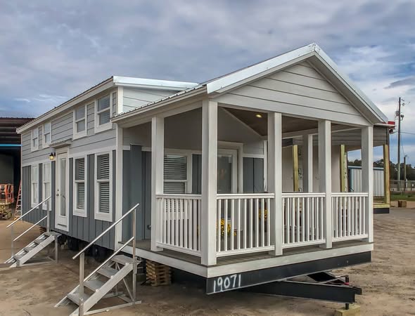 Compact grey tiny house on a platform with white trim. It has a cozy front porch and small staircases on each side. The sky is overcast.