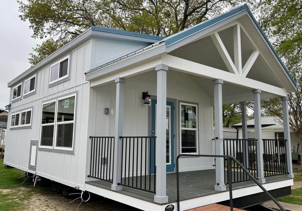 A modern tiny house with white siding and blue accents stands in a green, leafy area. It features large windows and a small porch with black railings.