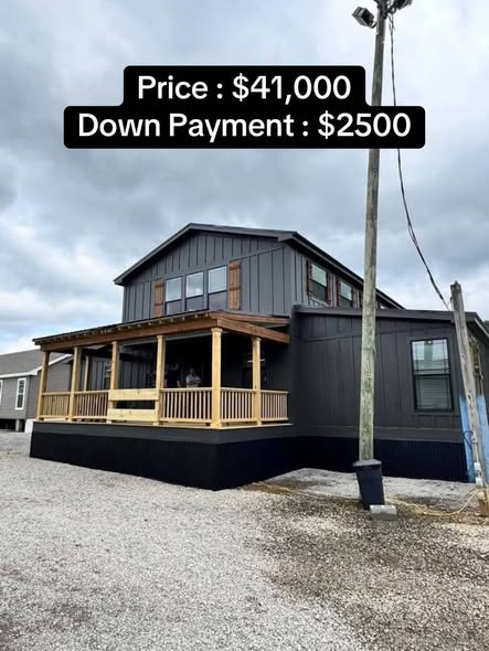 A modern, dark gray two-story house with wooden accents and a spacious porch sits under a cloudy sky. Price and down payment details are displayed above.