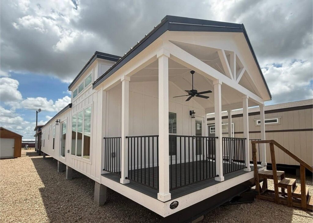 Modern tiny home exterior with a covered front porch featuring a ceiling fan and black railing. The home has large windows and a gabled roof under a partly cloudy sky.