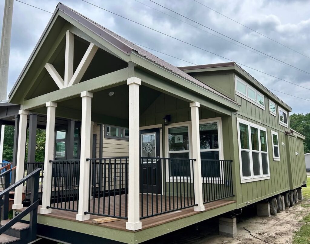 A green tiny house with a peaked roof and white trim sits on wheels. It features a covered porch with railings and large windows, under a cloudy sky.