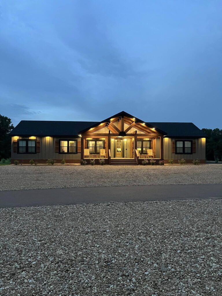A warmly lit modern farmhouse with a gabled roof at dusk, large front porch, and gravel driveway. The sky is overcast, enhancing the cozy ambiance.