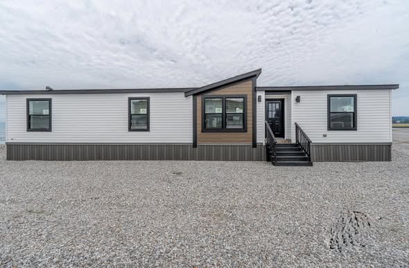 A modern, single-story manufactured home with a white facade and dark trim sits on a gravel lot. It features a small front porch and several windows.