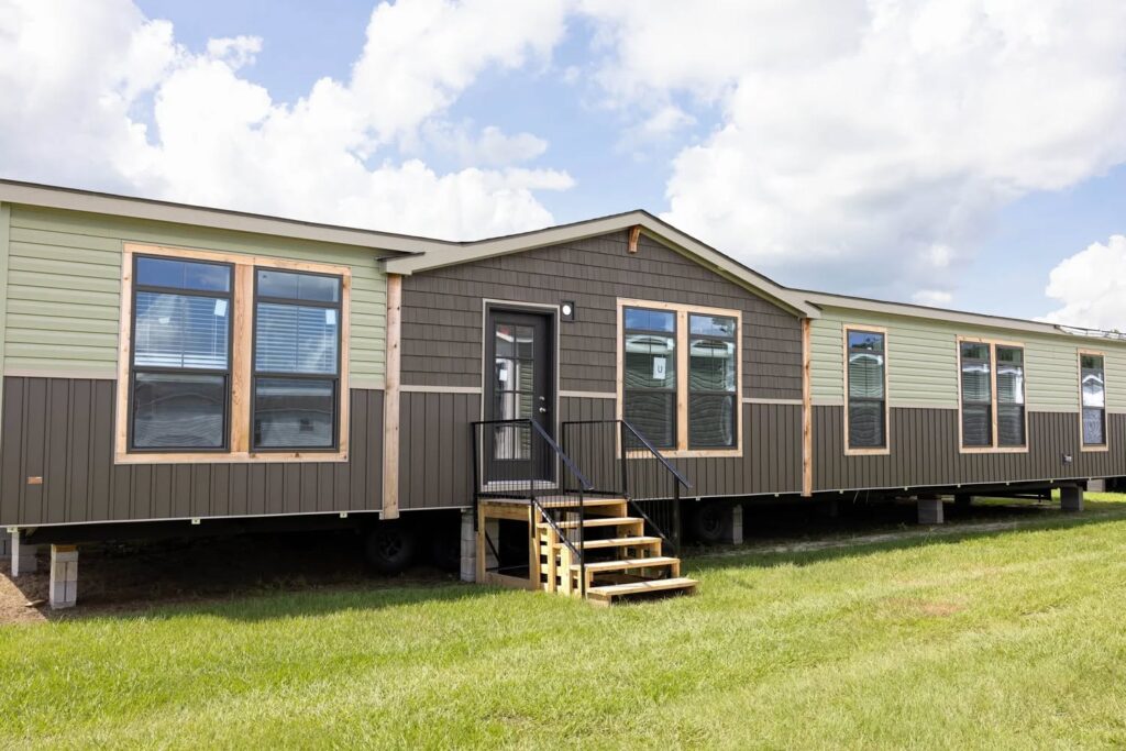 A modern manufactured home on a grassy lot under a blue sky with clouds. The home features large windows, light green and dark brown siding.