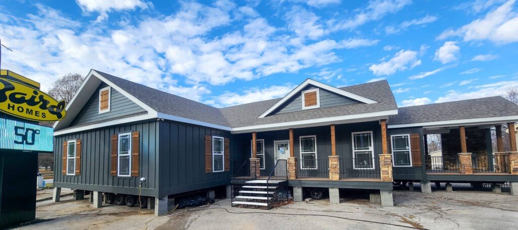 A modern, dark gray modular home with white trim and wooden accents sits on a lot under a blue sky. A sign on the left reads "ScotBilt Homes."