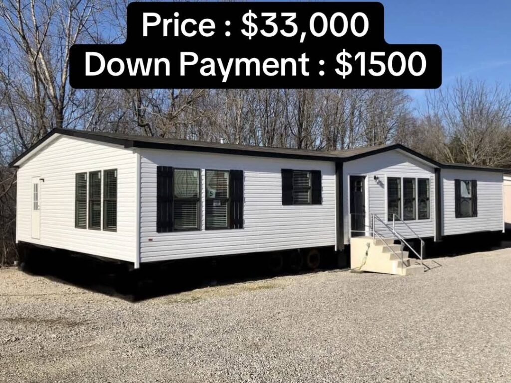 A white mobile home with black shutters is set on a gravel lot, surrounded by bare trees. Text above lists price as $33,000 and down payment as $1,500.