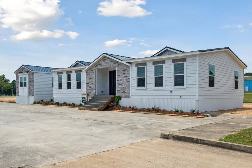 A modern white modular home with a gray gabled roof, stone accents, and multiple windows, set on a concrete driveway with landscaped edges under a blue sky.
