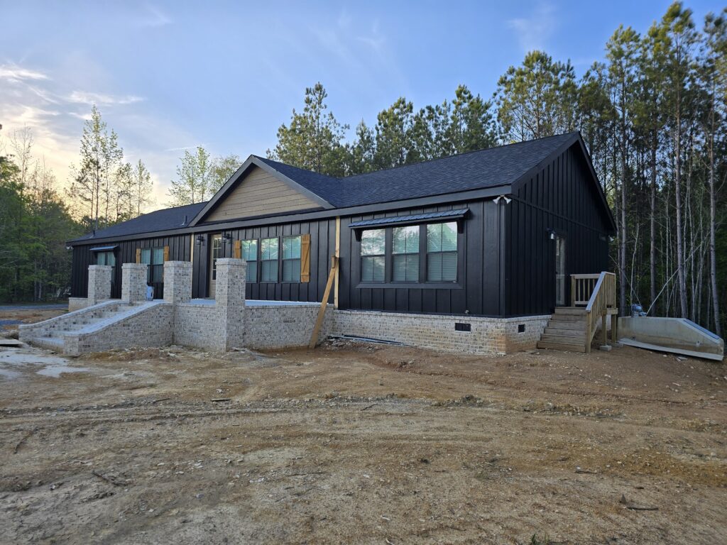 Single-story black house with brick foundation amid a wooded area. It has multiple windows, two staircases, and a clear blue sky overhead.