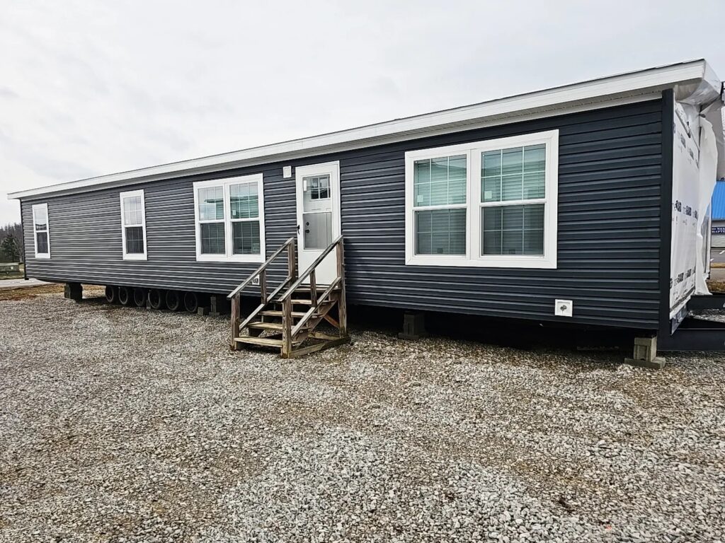 A modern, dark-colored mobile home with multiple large windows sits on a gravel plot. Wooden steps lead to the door, suggesting a simple yet functional design.