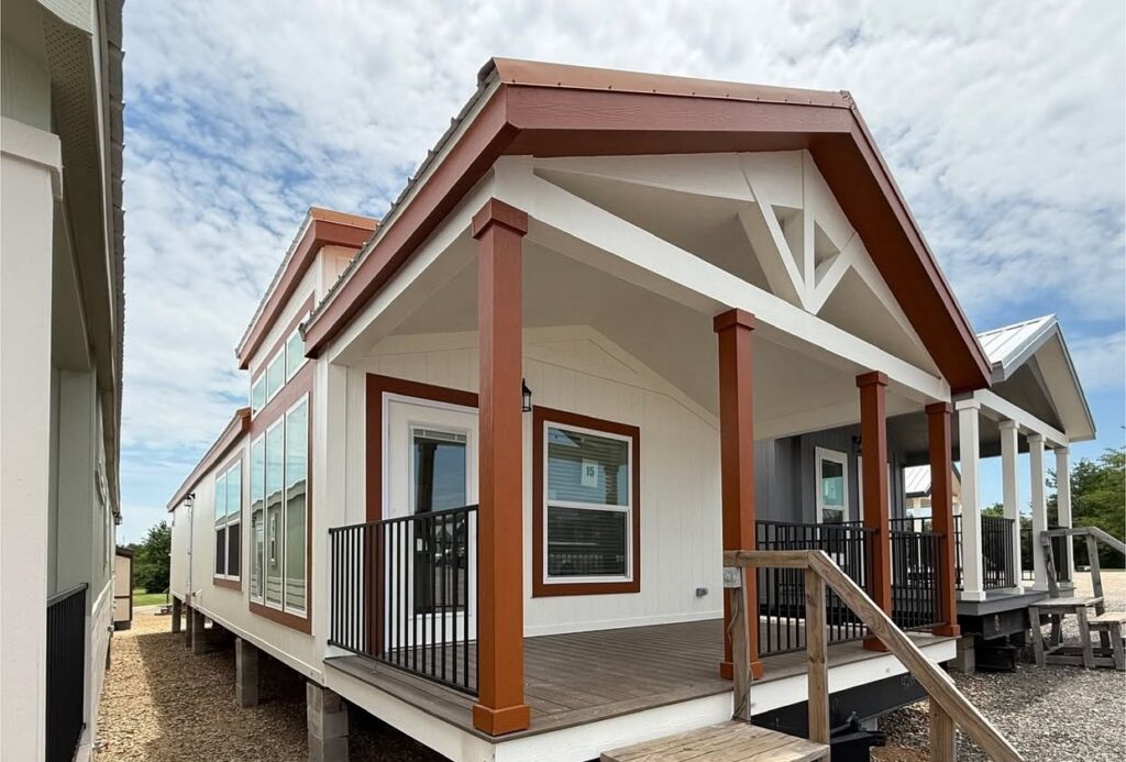A small, modern prefab house with white walls and red-brown trim features a front porch with black railings, set against a cloudy sky backdrop.