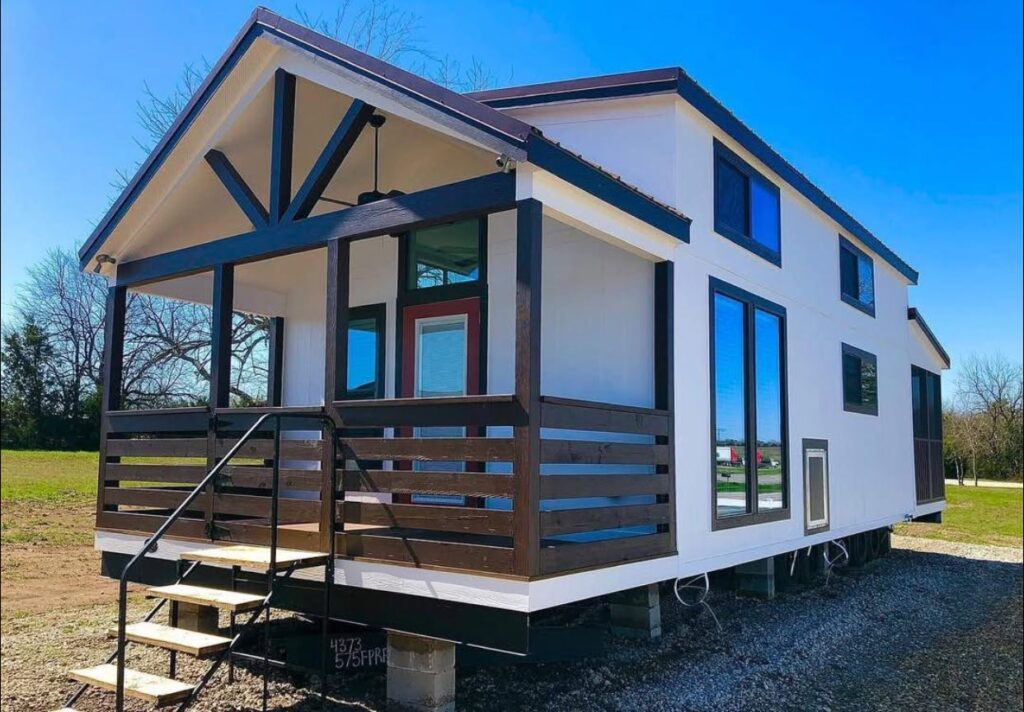 Modern tiny house with white walls and dark trim, featuring a front porch, large windows, and set on concrete blocks in a grassy area with bright blue sky.
