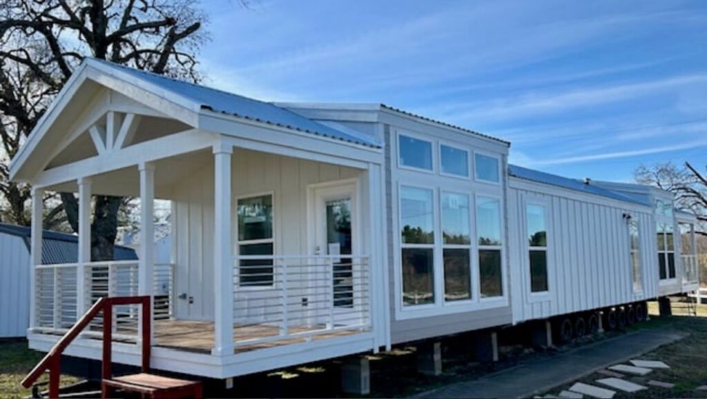 A modern white prefab home with large windows and a small front porch under a blue sky. The setting is calm, surrounded by trees and open space.
