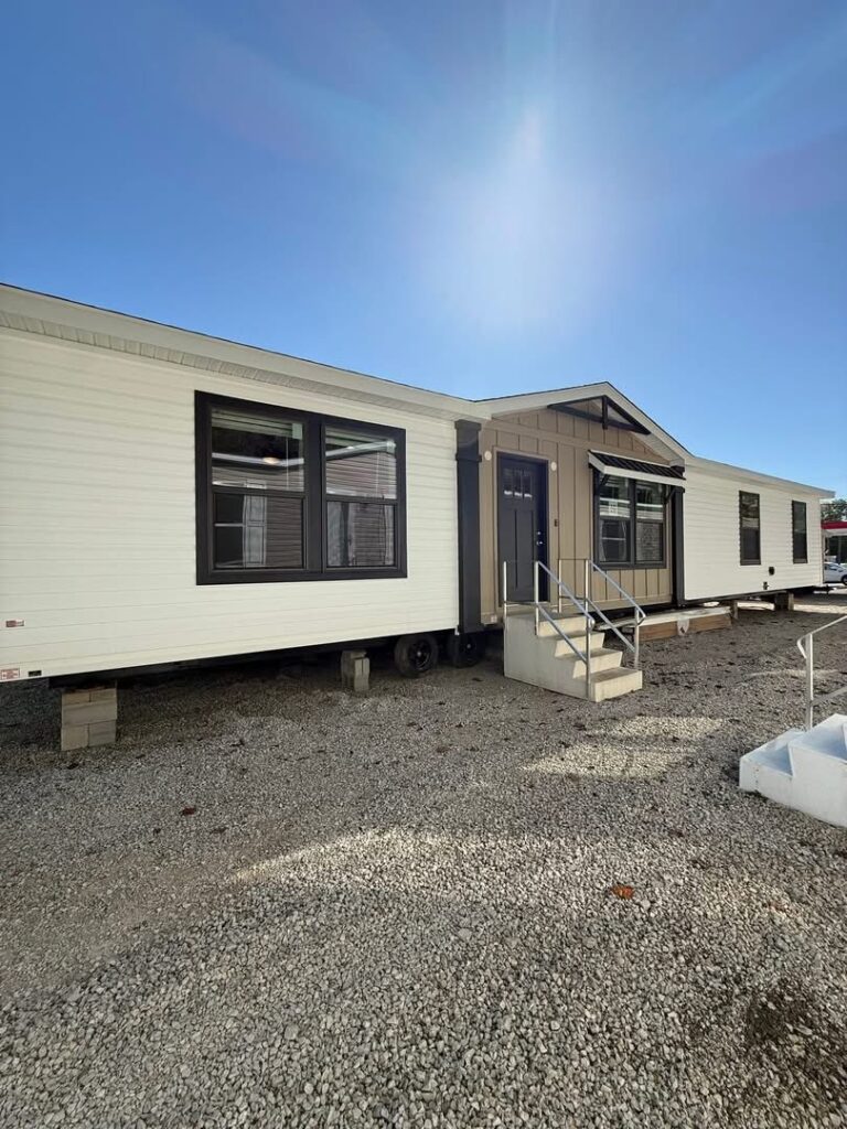 A large manufactured home with white siding and dark trim is raised on blocks against a clear blue sky. Sunlight casts soft shadows on the gravel ground.