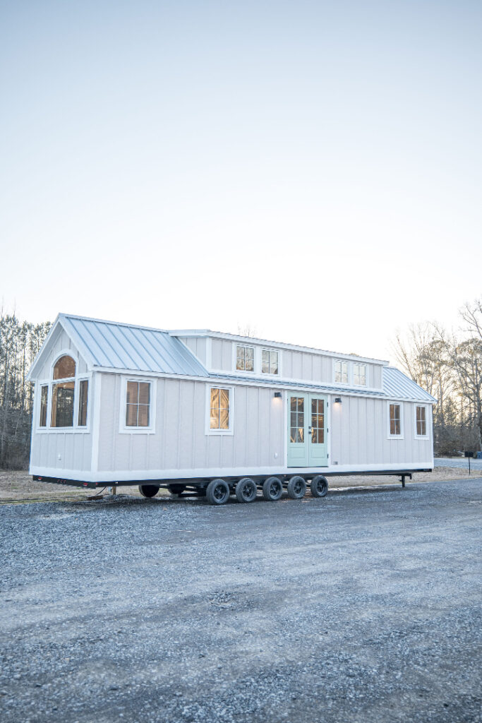 A white tiny house on wheels sits on a gravel path, featuring multiple windows and a small porch. The setting is serene, with a pale, early evening sky.