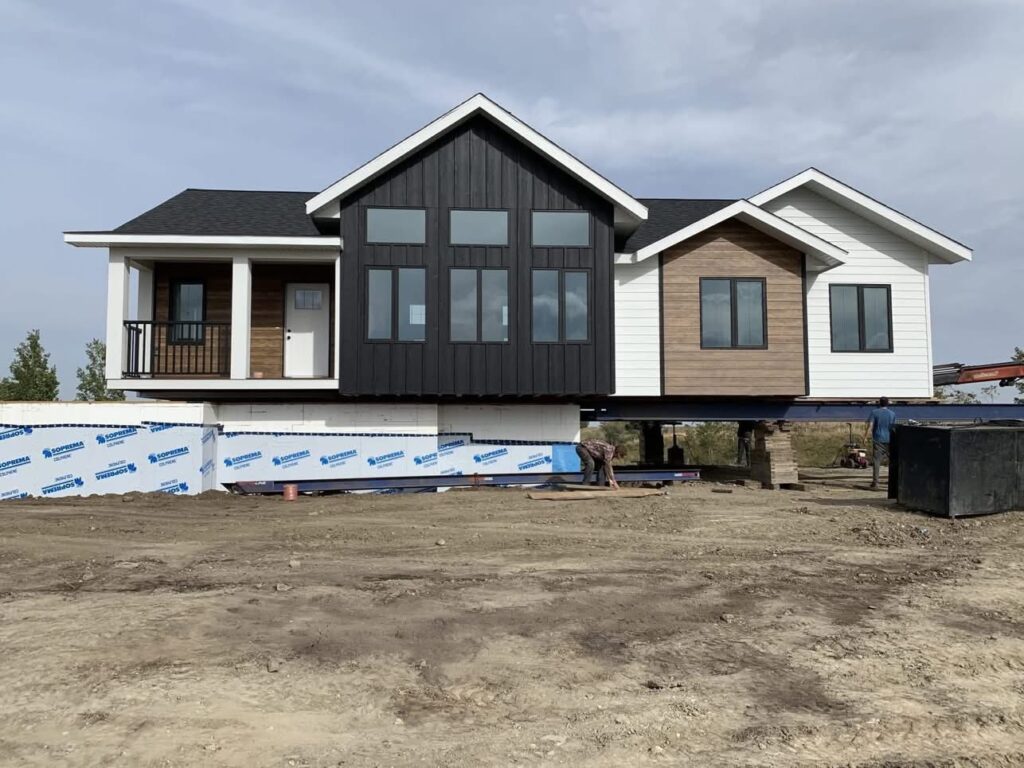 Modern modular house under construction, elevated on a foundation. Features a blend of dark, wood, and white panels, with large windows.