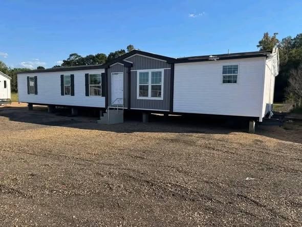A modern white modular home with a dark trim sits on a gravel lot against a backdrop of trees under a clear blue sky, conveying a sense of tranquility.