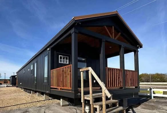 Modern tiny house with black siding and wood accents, elevated on a platform with stairs leading to a porch, set under a clear blue sky.